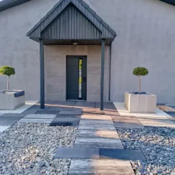 Nowoczesne wejście do domu z szarej kostki, żwiru i płyt. Zadaszenie nad drzwiami, donice z drzewkami. Minimalistyczny design, symetryczny układ.