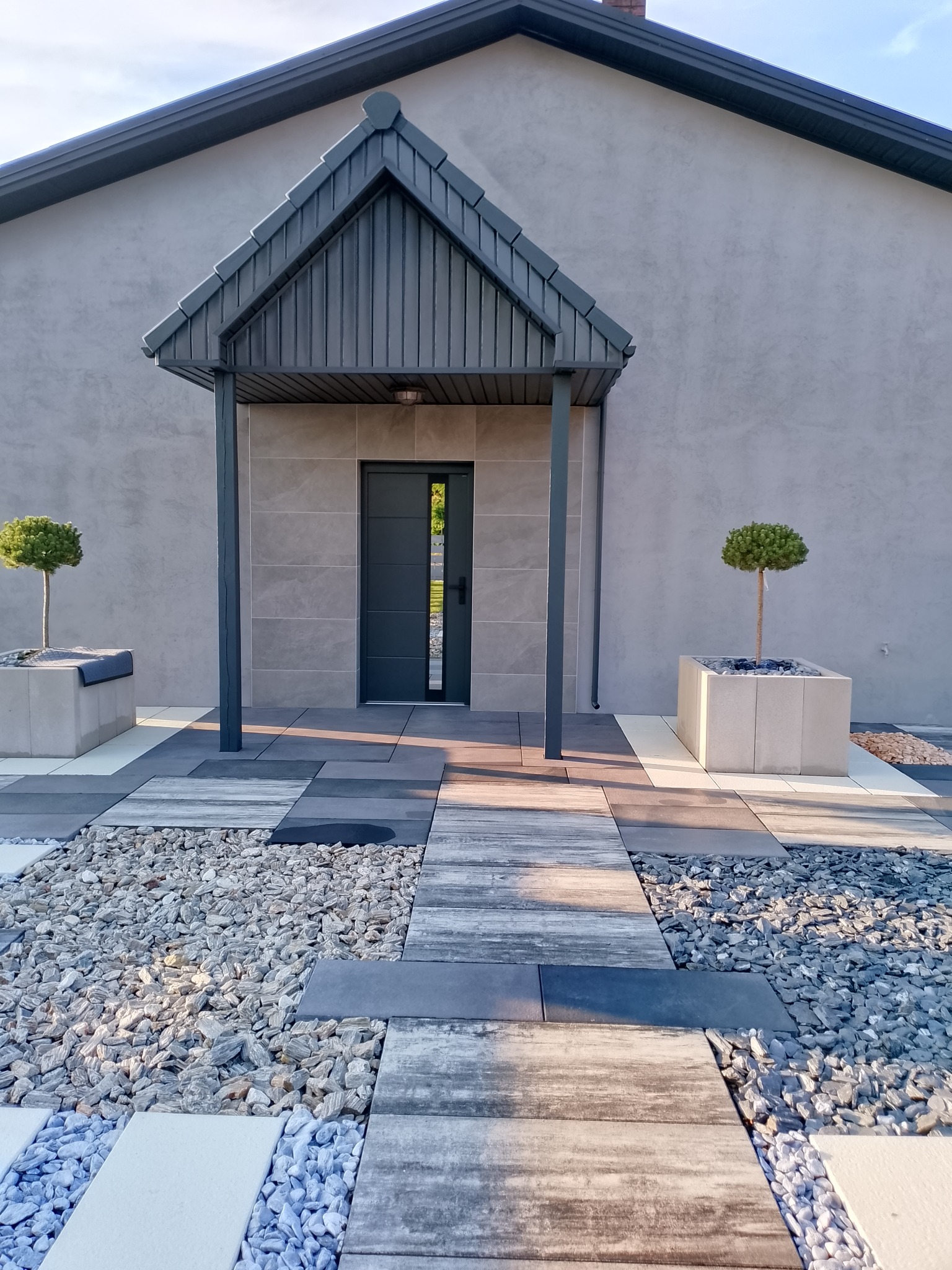 Nowoczesne wejście do domu z szarej kostki, żwiru i płyt. Zadaszenie nad drzwiami, donice z drzewkami. Minimalistyczny design, symetryczny układ.