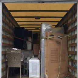 Wnętrze skrzyni ładunkowej wypełnione meblami ogrodowymi, zapakowanym materacem i kartonami zabezpieczonymi pasami transportowymi.