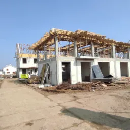 Budowa osiedla domów jednorodzinnych w stanie surowym otwartym, widoczne zbrojenia i szalunki stropowe, teren budowy z materiałami budowlanymi i przenośną toaletą.