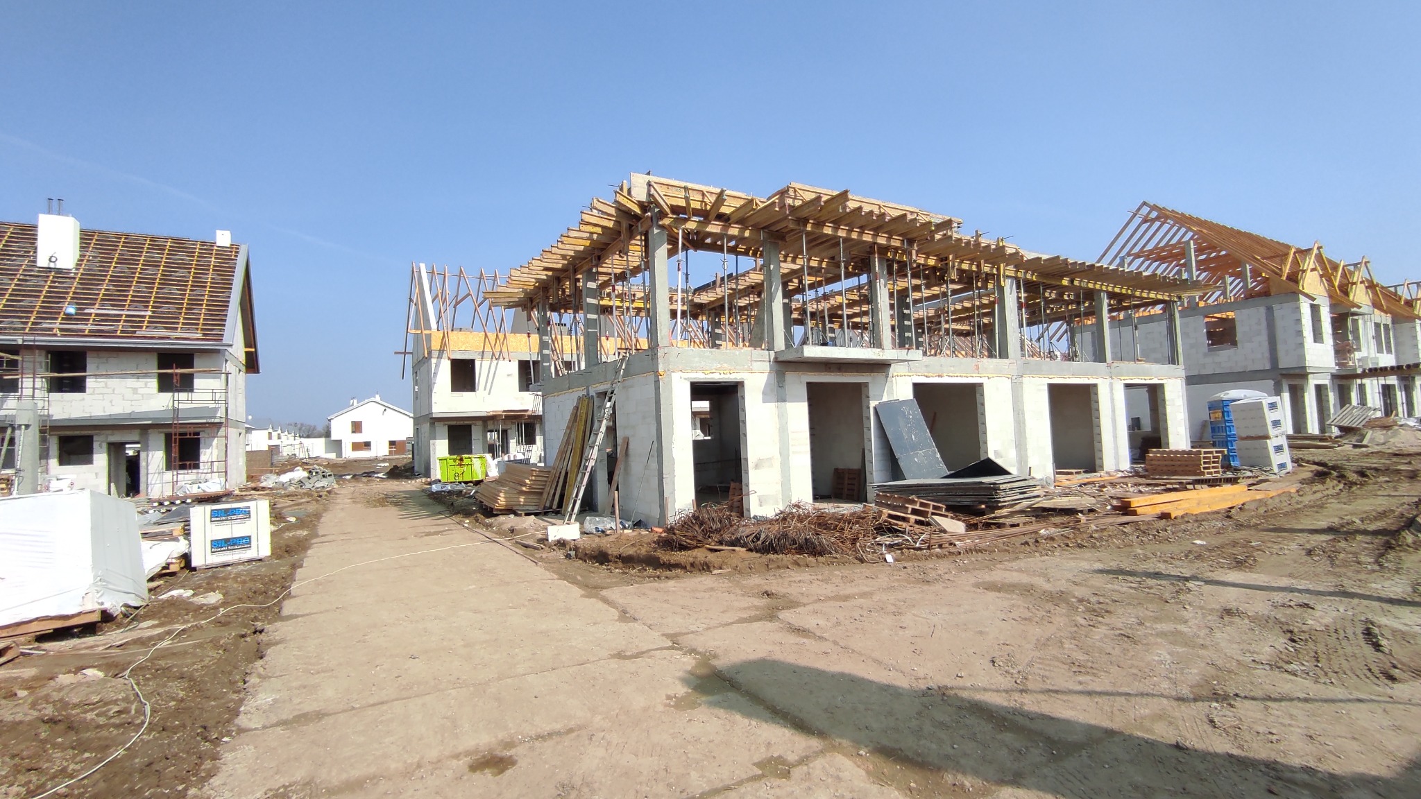 Budowa osiedla domów jednorodzinnych w stanie surowym otwartym, widoczne zbrojenia i szalunki stropowe, teren budowy z materiałami budowlanymi i przenośną toaletą.