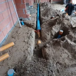 Wykop z ułożonym niebieskim rurociągiem kanalizacyjnym w surowym stanie budynku z cegły, widoczny pracownik w tle.