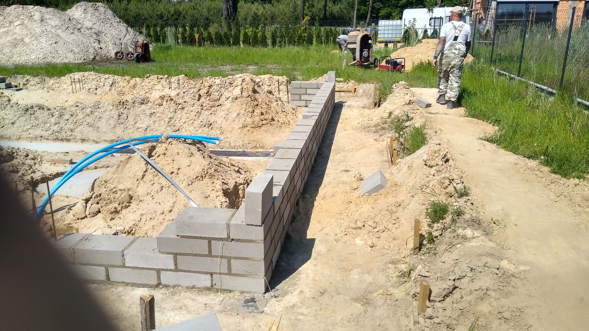 Budowa fundamentów z bloczków betonowych, widoczne niebieskie rury instalacyjne, koparka i pracownik w tle.