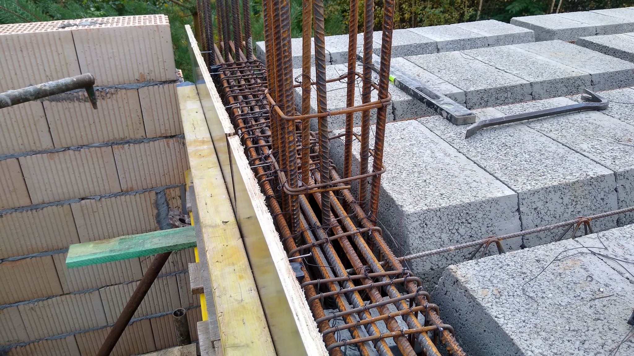 Wiązanie zbrojenia w szalunku fundamentowym z bloczków betonowych, widoczne poziomica i łom na górze, elementy podpór szalunku po lewej stronie