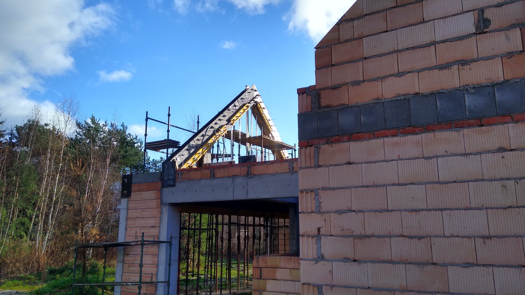 Wznoszenie ścian domu z bloczków ceramicznych, widoczne zbrojenie stropu i konstrukcja więźby dachowej w surowym stanie, na tle błękitnego nieba i drzew.