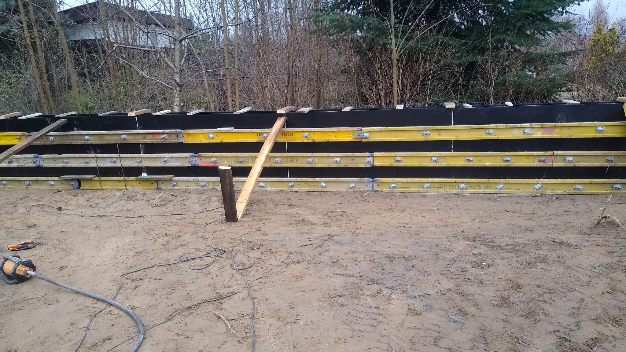 Szczegółowe ujęcie szalunku fundamentowego z żółtymi belkami H20 i czarną membraną, wspartego drewnianymi stemplami, na tle ziemi i drzew bez liści.