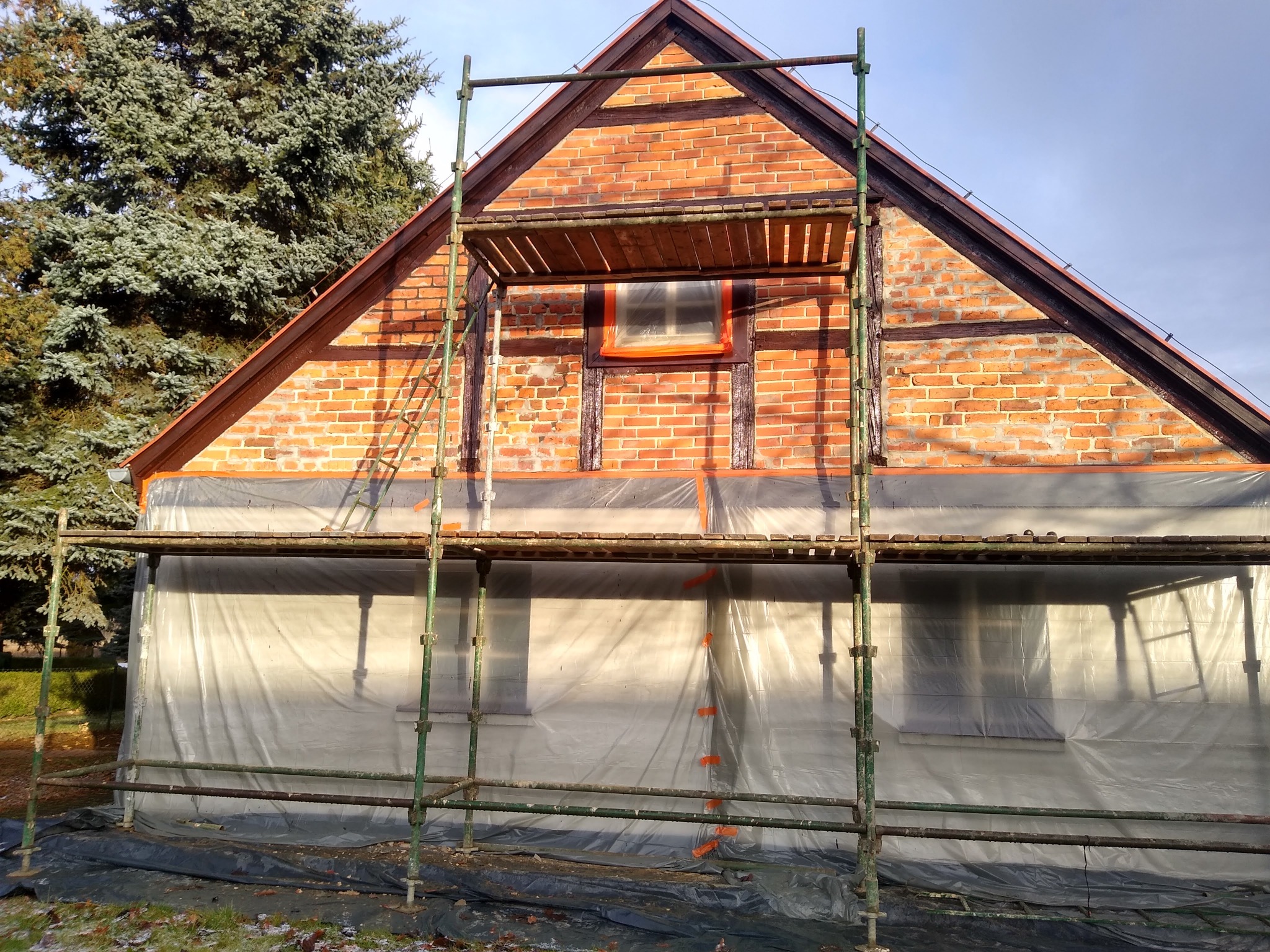 Dom z czerwonej cegły w trakcie prac elewacyjnych, osłonięty folią i otoczony rusztowaniem, widoczne elementy drewniane konstrukcji.