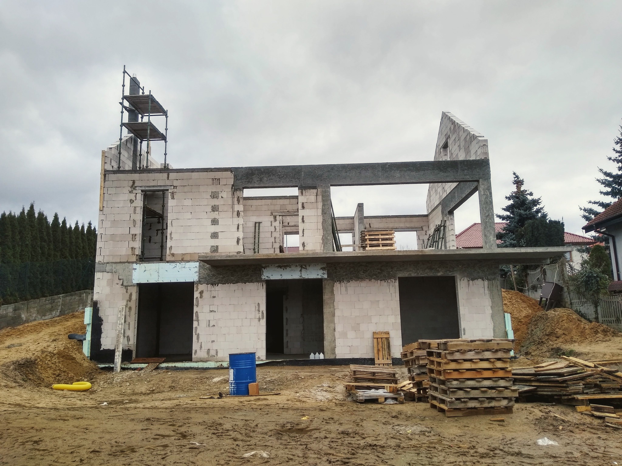 Niedokończony dwukondygnacyjny budynek z białego bloczka, częściowo ocieplony styropianem, z rusztowaniem, paletami i niebieską beczką na błotnistym placu budowy w pochmurny dzień.