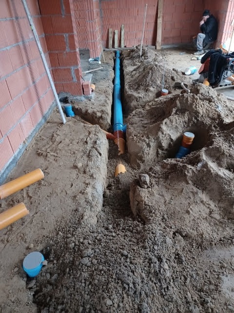Wykop z ułożonym niebieskim rurociągiem kanalizacyjnym w surowym stanie budynku z cegły, widoczny pracownik w tle.