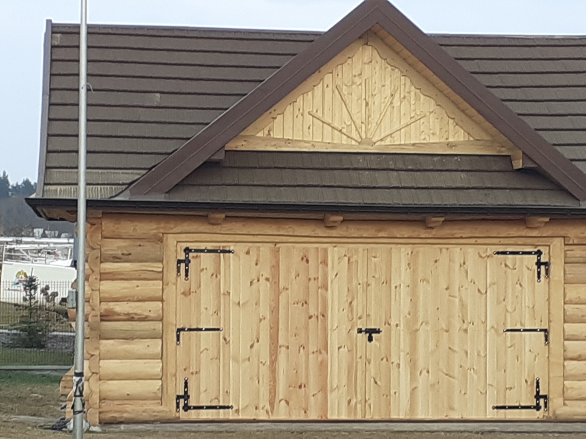 Garaż z bali z dwuskrzydłowymi drewnianymi wrotami, czarnymi okuciami i brązowym dachem z drewnopodobnej dachówki, ozdobny szczyt w formie słońca.