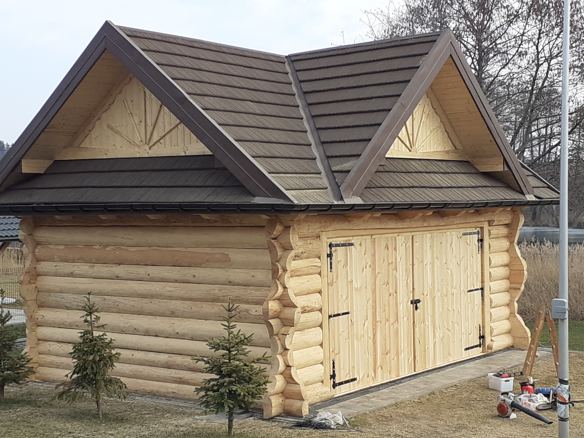 Nowoczesny dom z bali z dwuskrzydłowymi drzwiami garażowymi, pokryty brązową dachówką bitumiczną, z widocznymi narzędziami i małą choinką przed wejściem.