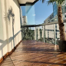 Nowoczesny balkon z drewnianą podłogą, przeszkloną balustradą i palmą w donicy, cień palmy na ścianie.