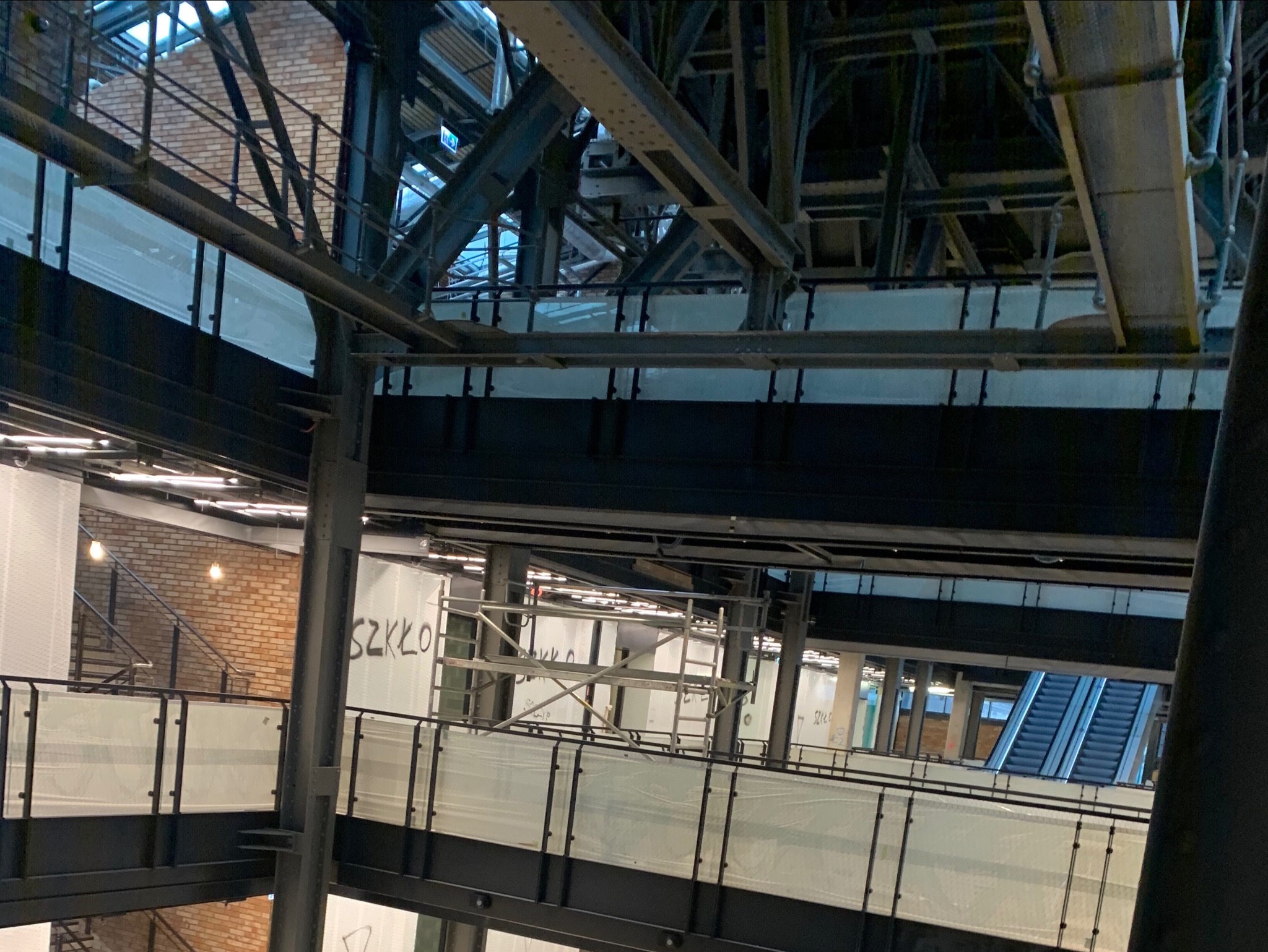 Wnętrze budynku w trakcie prac wykończeniowych, widoczne metalowe konstrukcje, rusztowania, szklane balustrady oraz fragment schodów ruchomych.