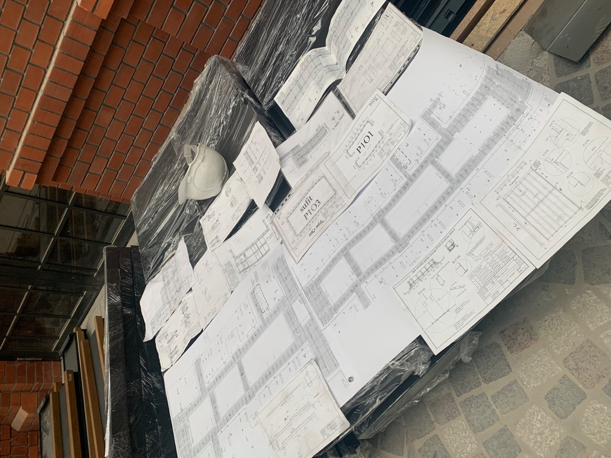 Stos planów architektonicznych z detalami technicznymi i biały kask ochronny leżące na palecie owiniętej folią stretch, obok ceglanej ściany.