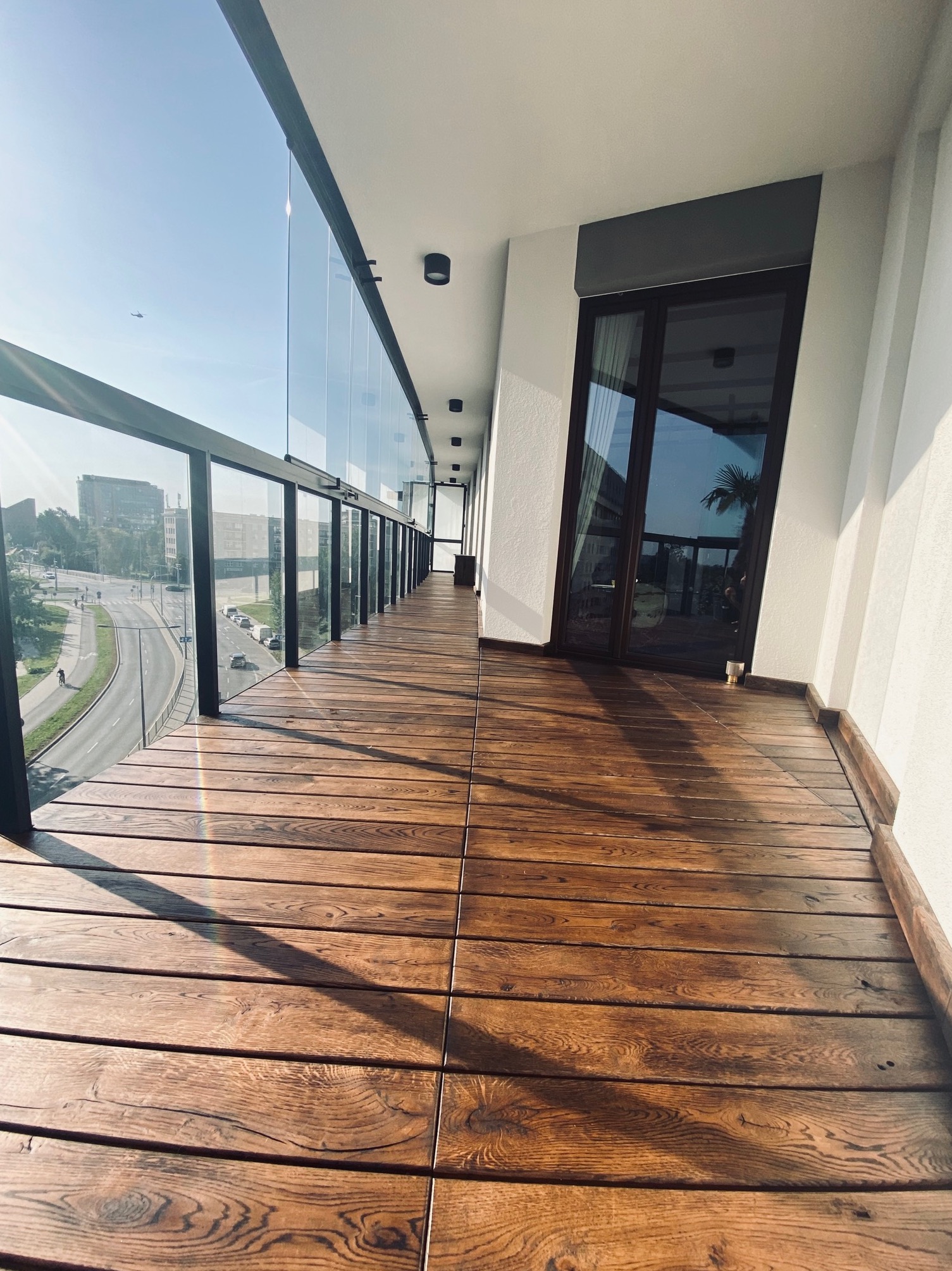 Długi balkon z drewnianą podłogą ułożoną z desek, szklana balustrada z czarnymi elementami konstrukcyjnymi, widok na miasto w tle, w górze widoczny samolot.
