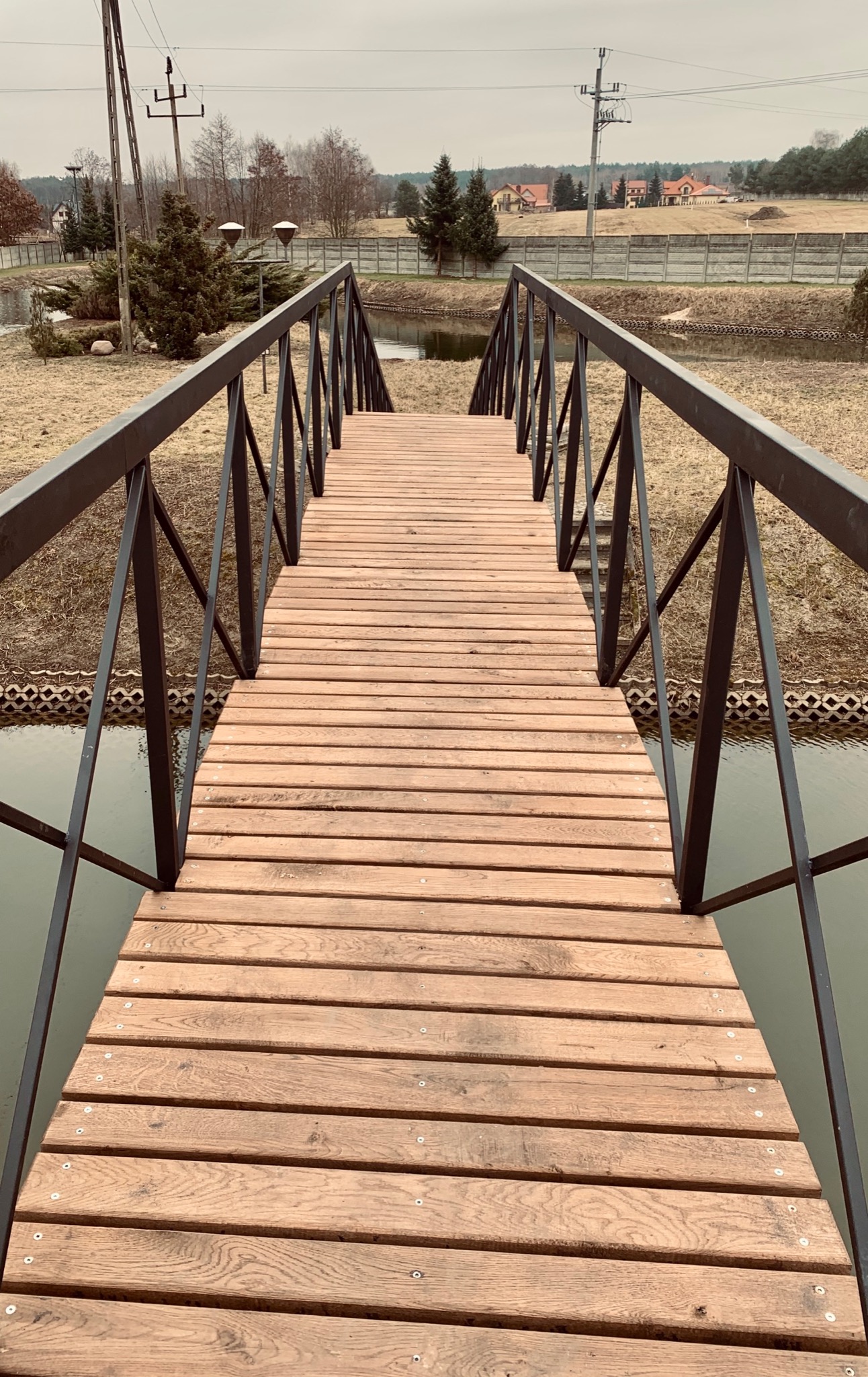 Drewniany mostek z czarną metalową balustradą nad niewielkim stawem, widok z perspektywy pieszego, w tle domy jednorodzinne i drzewa.