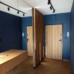 Nowoczesny przedpokój z granatowymi ścianami, drewnianą podłogą ułożoną w jodełkę, lustrem na ścianie i podświetlaną ścianką działową z drewnianych listew.