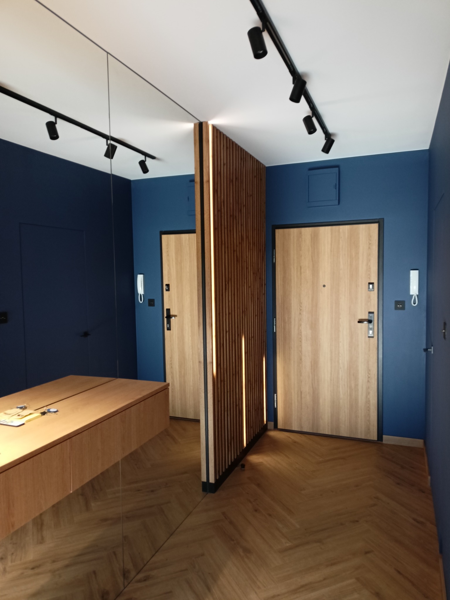 Nowoczesny przedpokój z granatowymi ścianami, drewnianą podłogą ułożoną w jodełkę, lustrem na ścianie i podświetlaną ścianką działową z drewnianych listew.