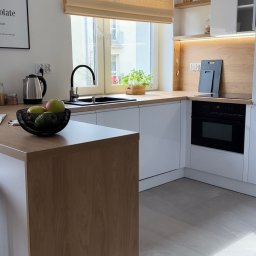 Arte Barbara Oleksa - Jasna kuchnia w stylu skandynawskim z drewnianym blatem, białymi szafkami i roletą w oknie. Na blacie misa z owocami i czajnik. Minimalistyczny design.