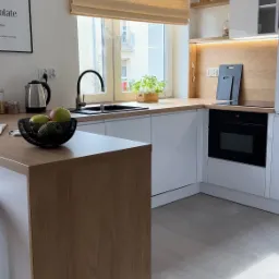 Jasna kuchnia w stylu skandynawskim z drewnianym blatem, białymi szafkami i roletą w oknie. Na blacie misa z owocami i czajnik. Minimalistyczny design.