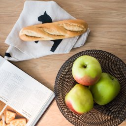 Arte Barbara Oleksa - Baguette na serwetce z motywem, jabłka w drucianym koszu i otwarta książka kucharska na drewnianym stole. Jasne, naturalne oświetlenie.