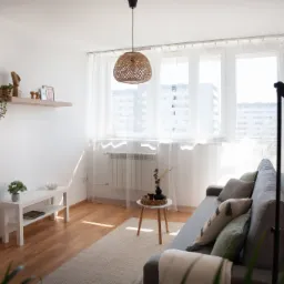 Jasne wnętrze salonu z szarą sofą, białymi ścianami i drewnianą podłogą. W oknie firanki, a nad sofą wisi lampa z abażurem z wikliny. Dekoracje na półkach i stoliku.