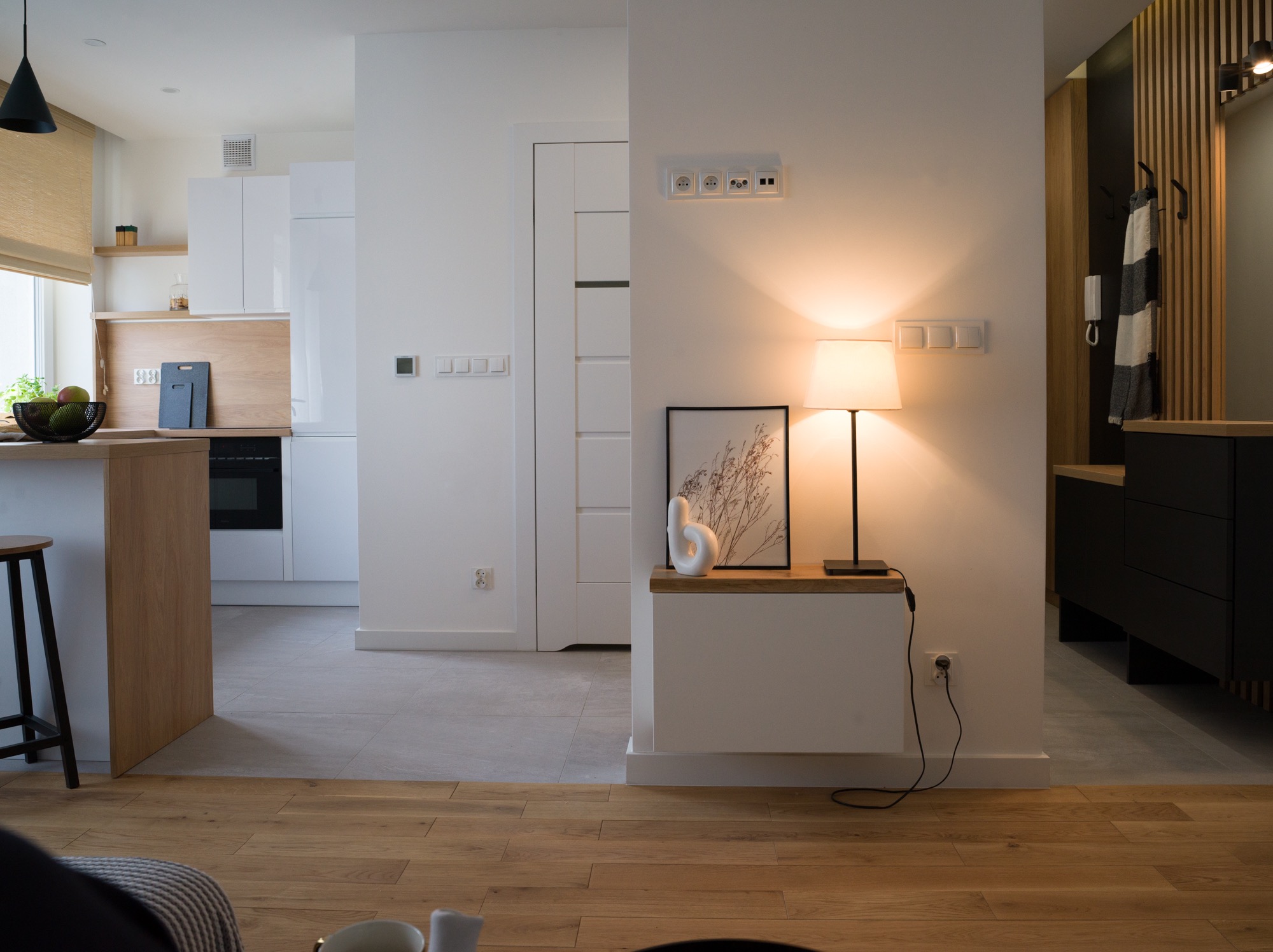 Nowoczesne wnętrze: jasna kuchnia z drewnianym blatem, minimalistyczne meble, lampa stołowa i dekoracje. Widok z salonu na otwartą przestrzeń.