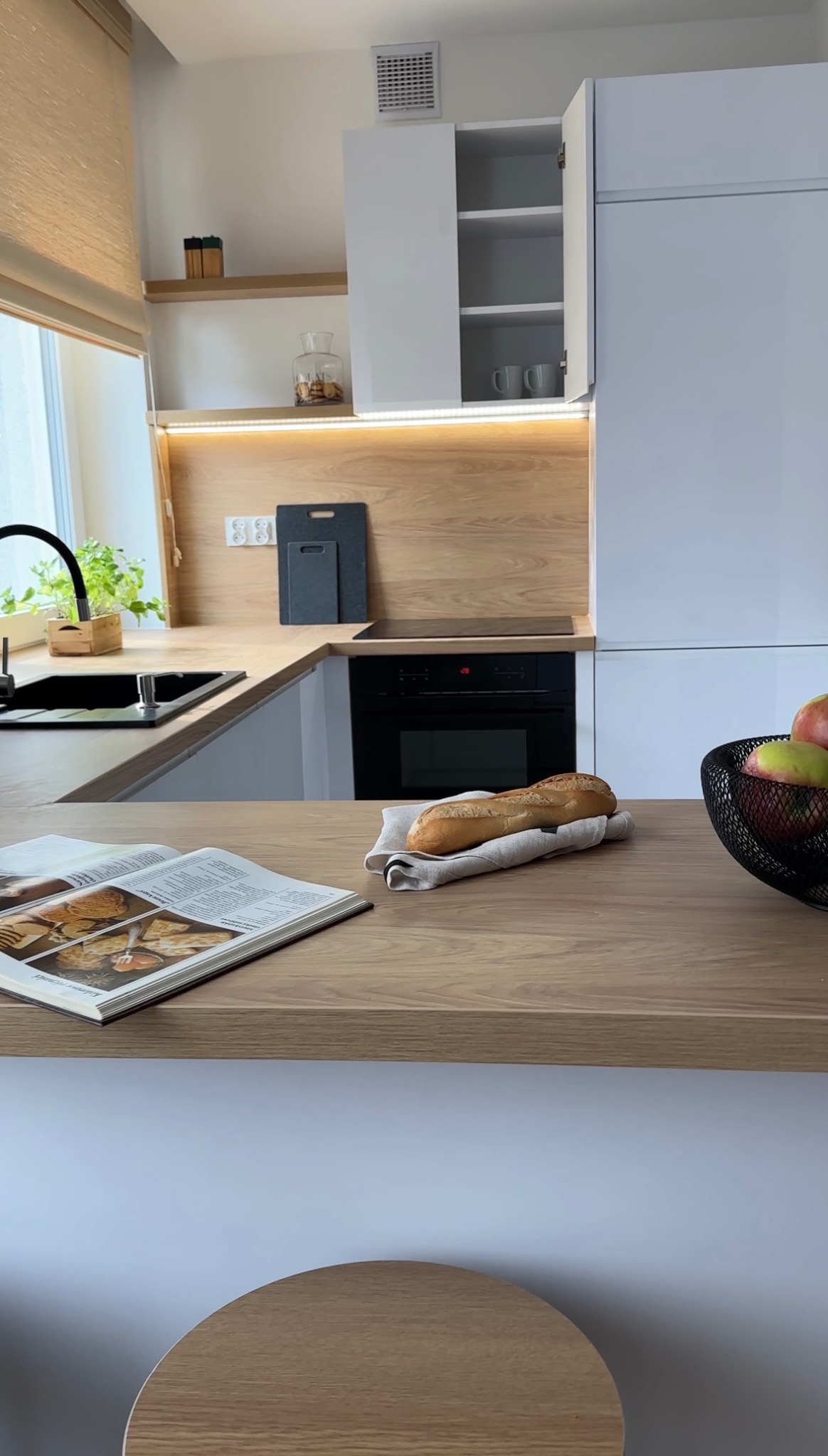 Jasna kuchnia z drewnianymi akcentami w Chorzowie: minimalistyczne szafki, drewniany blat z książką kucharską i bagietką, owocowy kosz. Nowoczesny design.