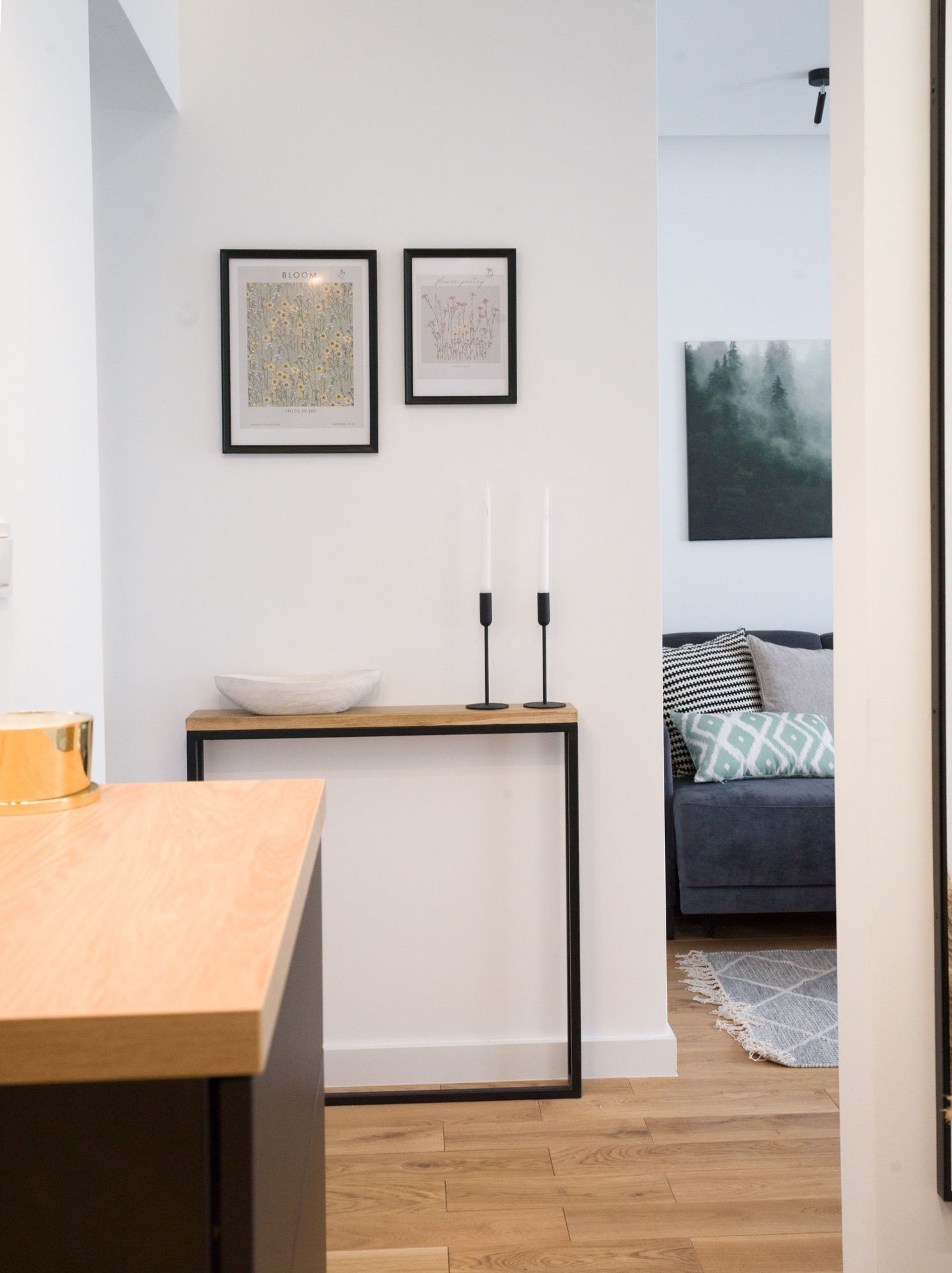 Nowoczesny przedpokój z drewnianą podłogą, konsolą z dekoracjami i widokiem na salon z szarą sofą i wzorzystymi poduszkami. Minimalistyczny design.