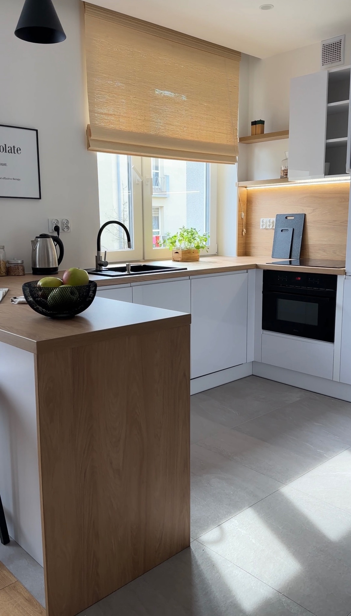 Jasna kuchnia w stylu skandynawskim z drewnianym blatem, białymi szafkami i roletą w oknie. Na blacie misa z owocami i czajnik. Minimalistyczny design.