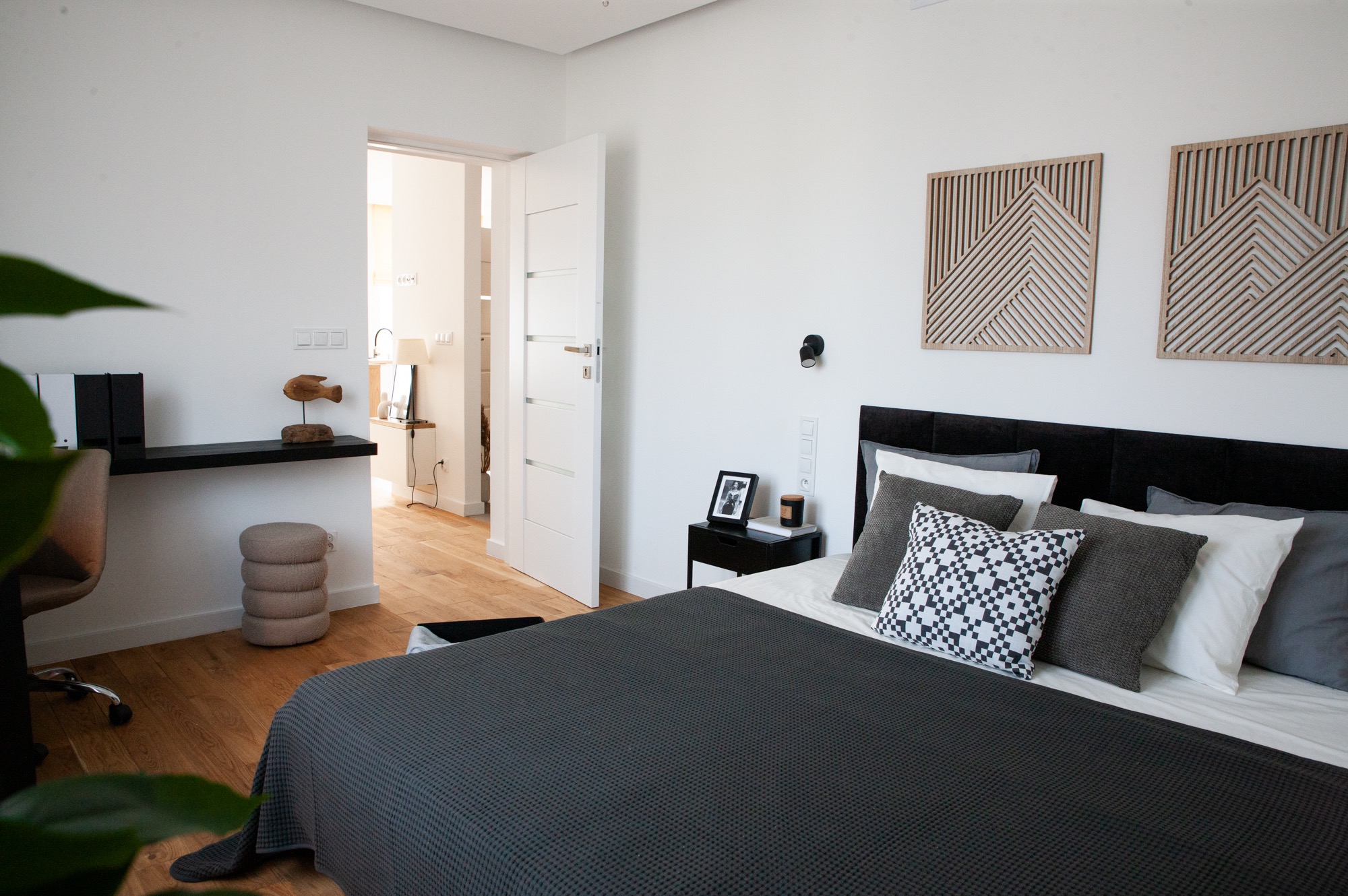 Nowoczesna sypialnia z drewnianą podłogą, czarnym zagłówkiem łóżka i dekoracjami ściennymi. Widok na otwarte drzwi do innego pomieszczenia.