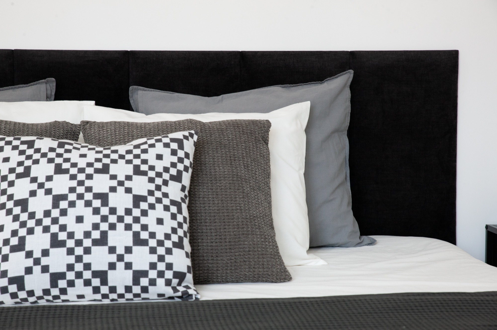 Minimalistyczna sypialnia: czarne wezgłowie, biała pościel i dekoracyjne poduszki w odcieniach szarości oraz wzorzysta poduszka w geometryczny deseń.