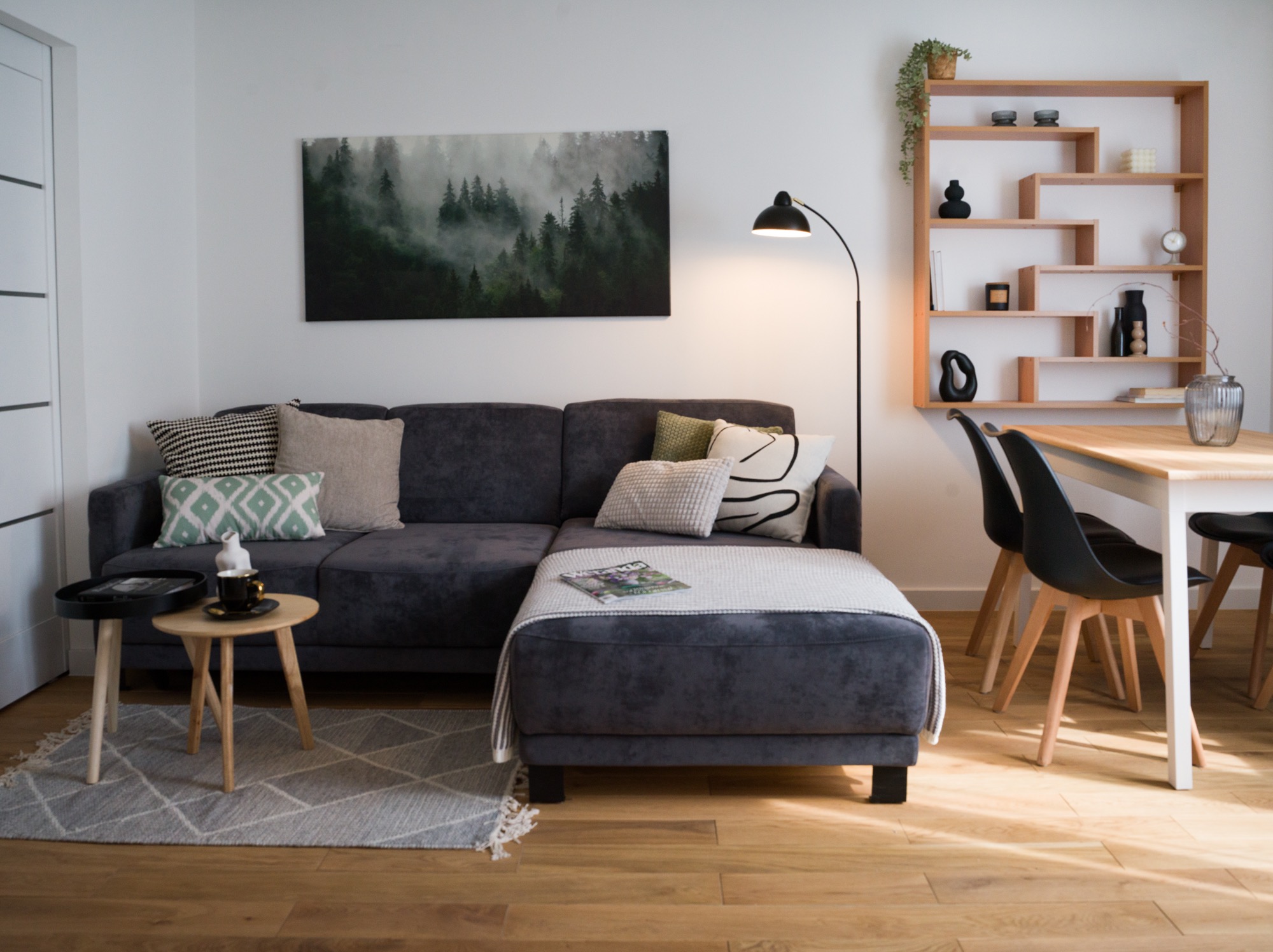 Nowoczesny salon z szarą sofą narożną, stolikami kawowymi i drewnianym regałem. Na ścianie obraz z motywem leśnym. Jasne drewniane podłogi i dywan w geometryczne wzory.