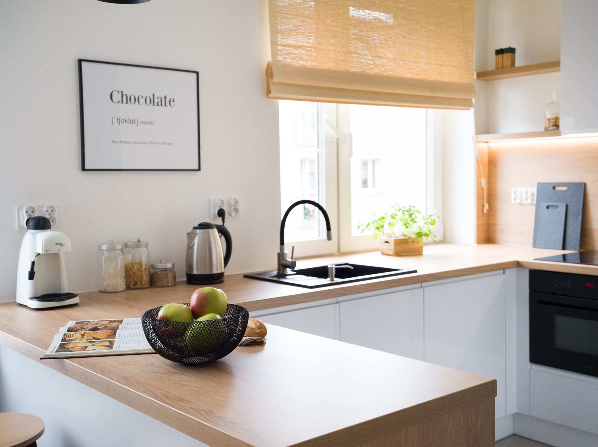 Jasna kuchnia z drewnianym blatem, na którym leżą owoce w metalowej misie i otwarta książka kucharska. W tle okno z roletą i plakat z napisem 'Chocolate'.