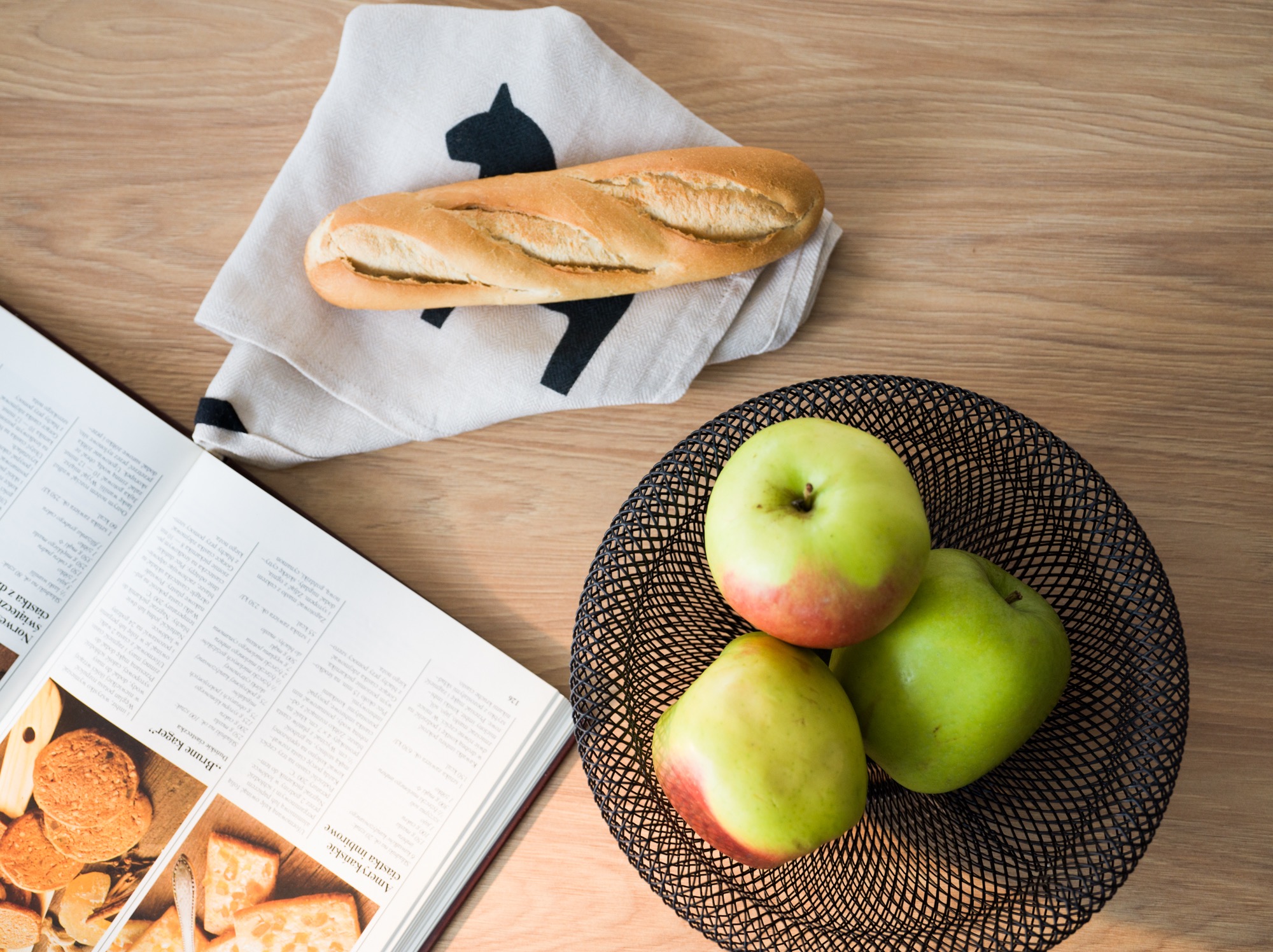 Baguette na serwetce z motywem, jabłka w drucianym koszu i otwarta książka kucharska na drewnianym stole. Jasne, naturalne oświetlenie.