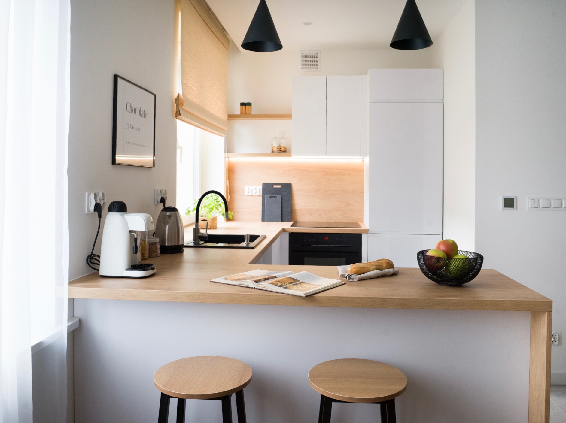 Jasna kuchnia w stylu skandynawskim z drewnianym blatem, czarnymi lampami i minimalistycznym wystrojem. Na blacie owoce, pieczywo i otwarta książka.