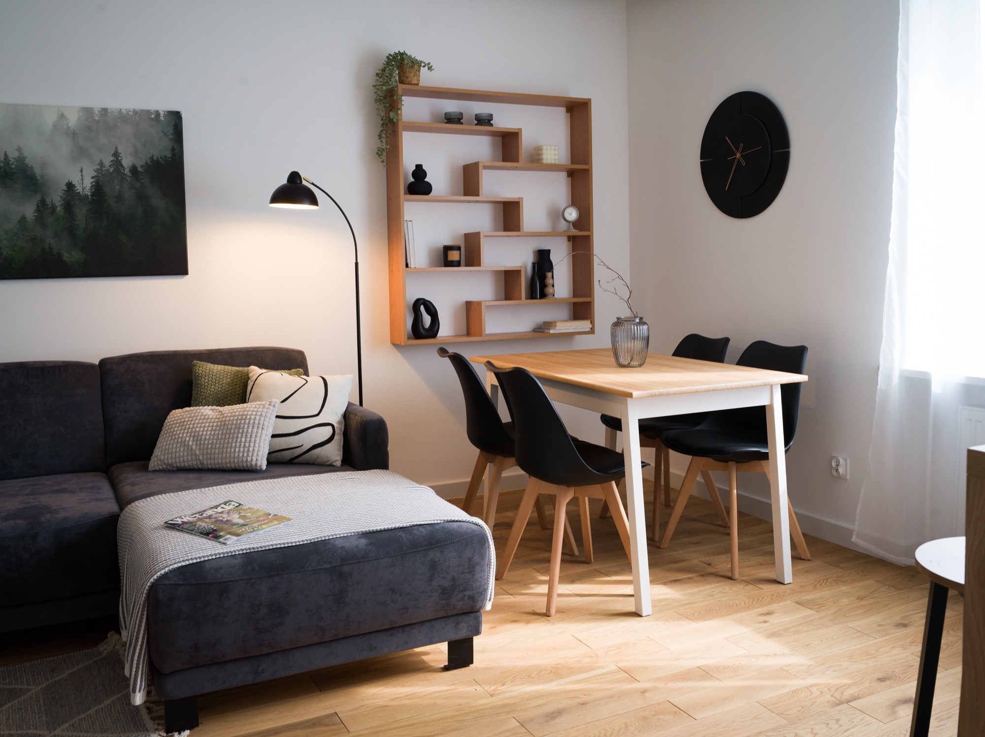 Jasne wnętrze: szara sofa z poduszkami, drewniany stół z czarnymi krzesłami, półka z dekoracjami i obraz lasu na ścianie. Minimalistyczny design.