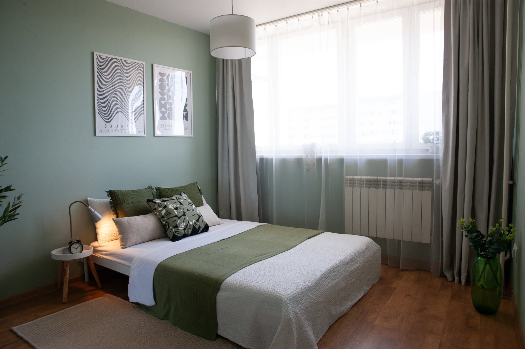 Jasna sypialnia z łóżkiem, zielonymi akcentami i plakatami Bauhaus na ścianie. Minimalistyczny wystrój z lampką nocną i drewnianą podłogą.