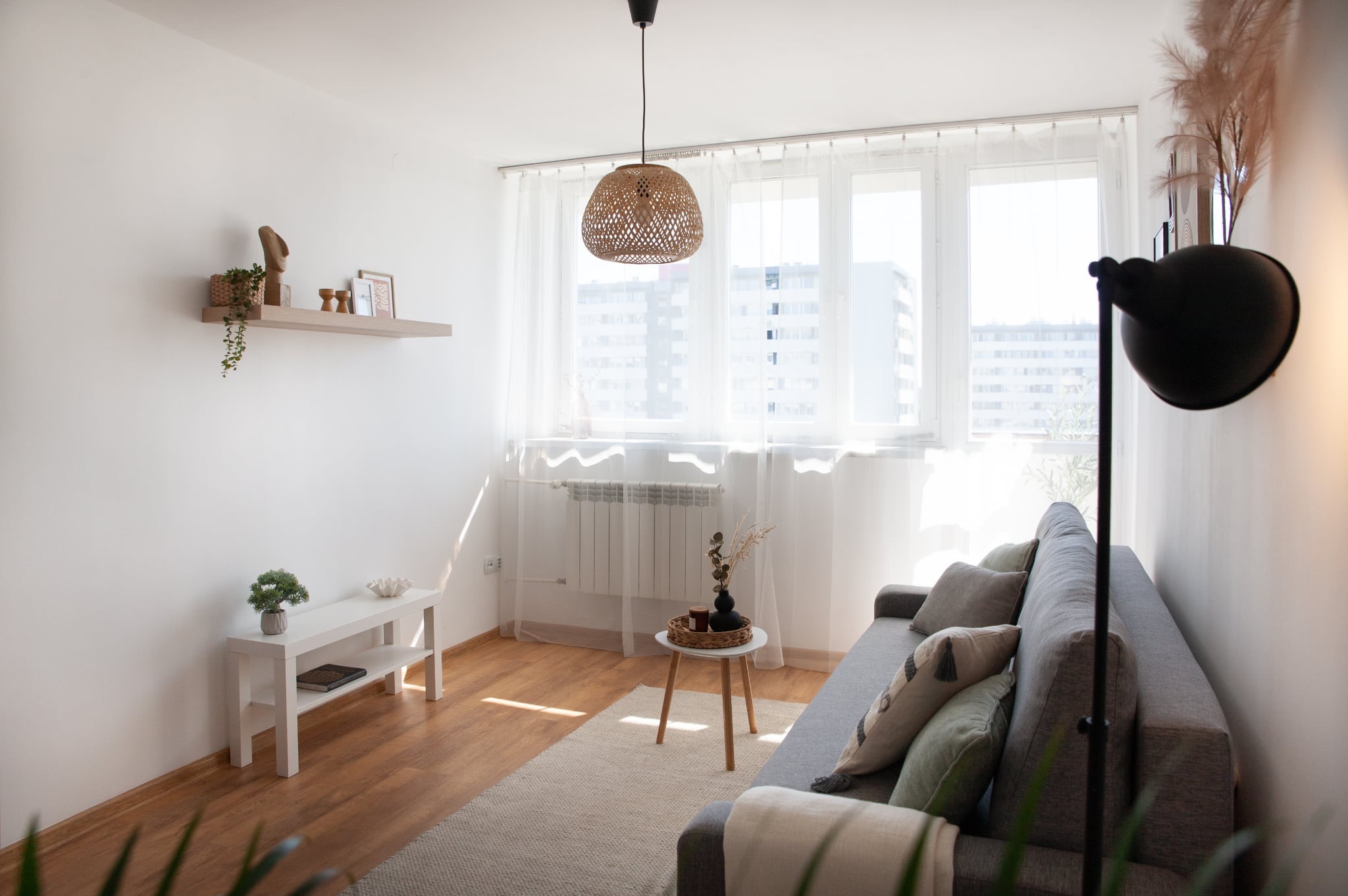 Jasne wnętrze salonu z szarą sofą, białymi ścianami i drewnianą podłogą. W oknie firanki, a nad sofą wisi lampa z abażurem z wikliny. Dekoracje na półkach i stoliku.