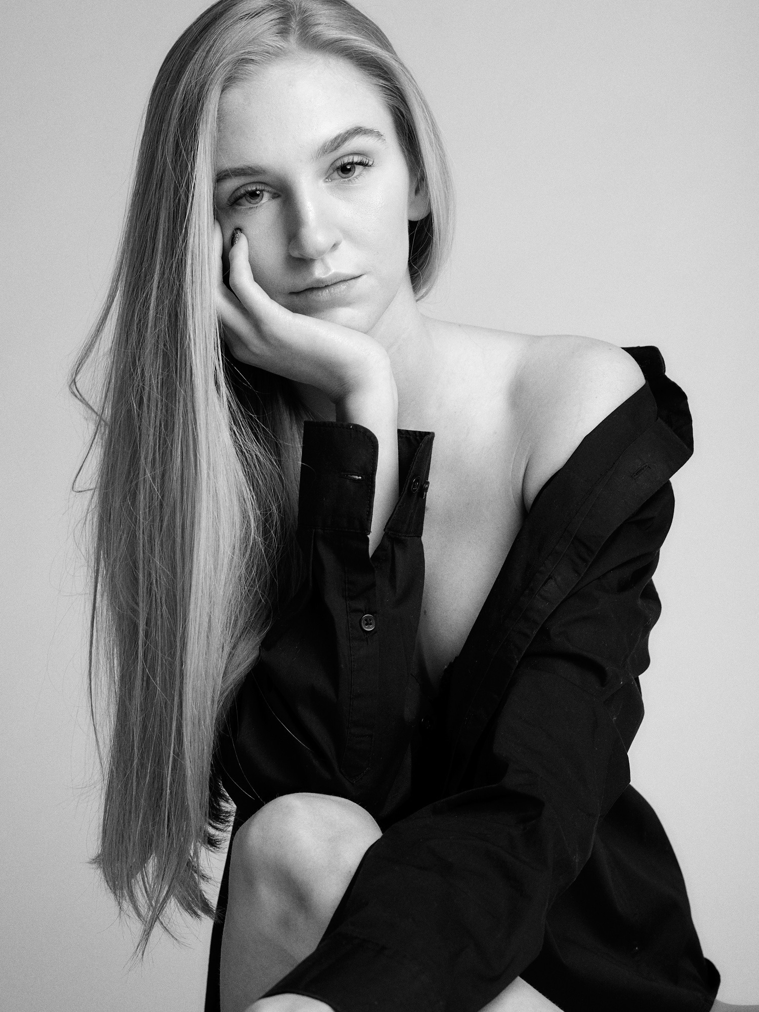 Portret kobiety z długimi blond włosami w czarnej, rozpiętej koszuli na neutralnym tle, w czerni i bieli.