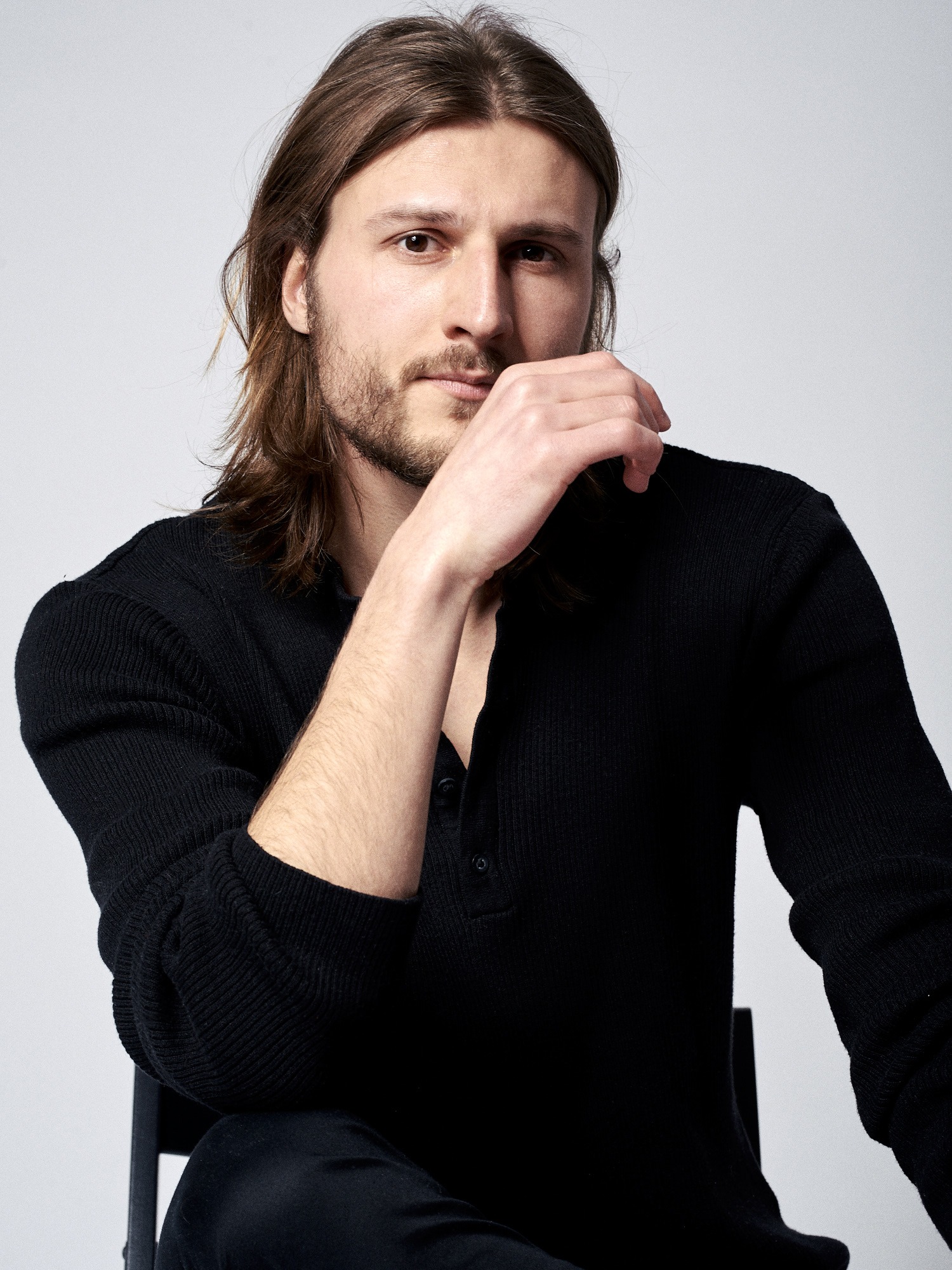 Portret mężczyzny z długimi włosami, w czarnej koszuli, siedzącego na krześle i dotykającego brody dłonią, na jasnym tle.