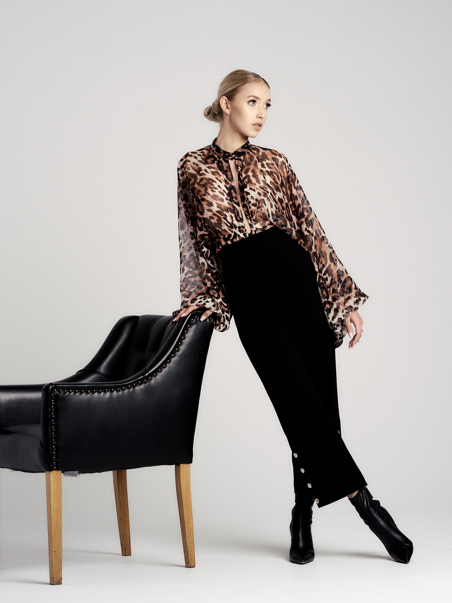 Kobieta w bluzce w panterkę i czarnej spódnicy pozuje opierając się o czarny skórzany fotel na drewnianych nogach, patrząc w bok.