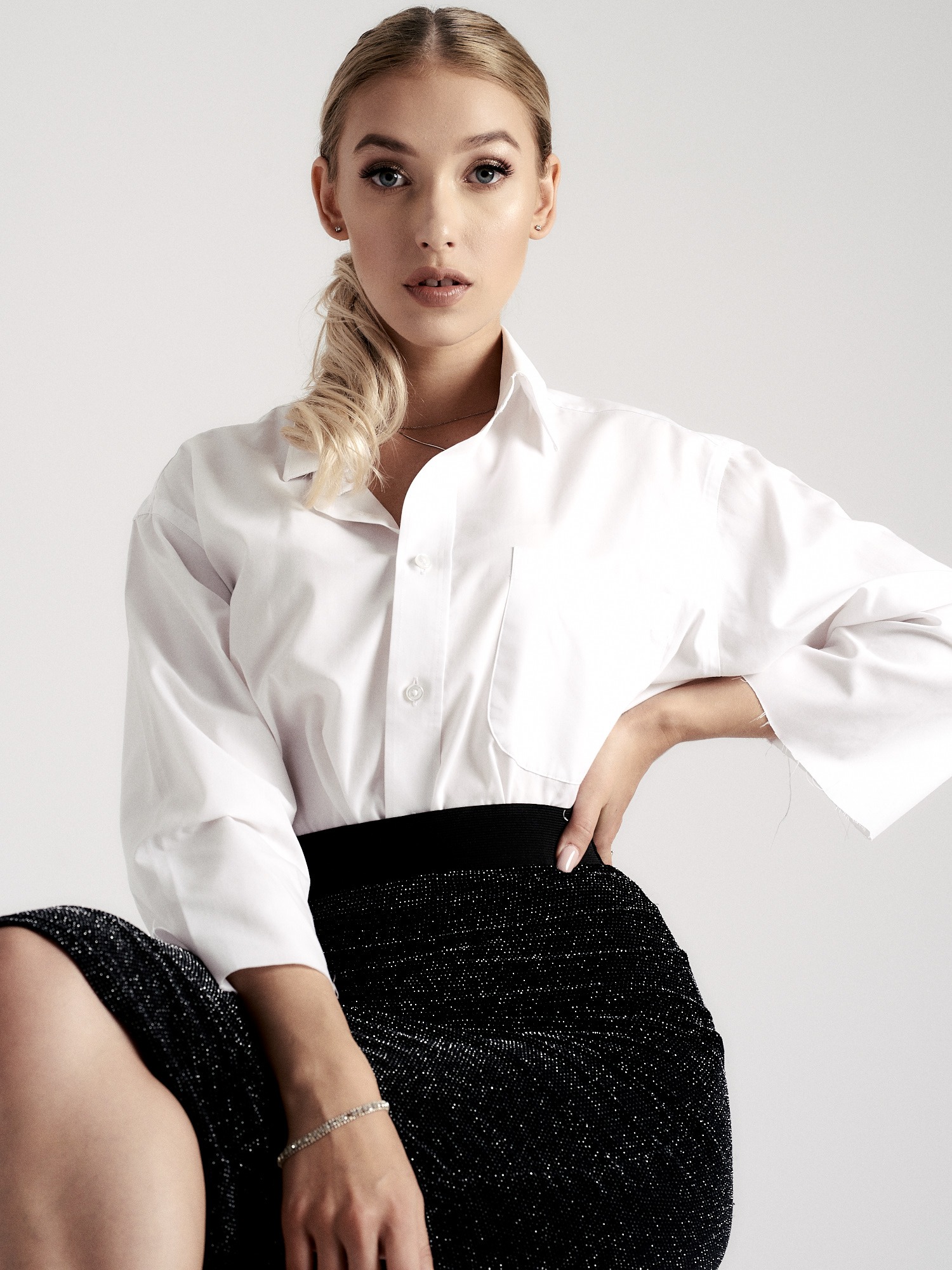 Portret kobiety w białej koszuli i czarnej, błyszczącej spódnicy, z włosami upiętymi w niski kucyk, pozującej na jasnym tle.