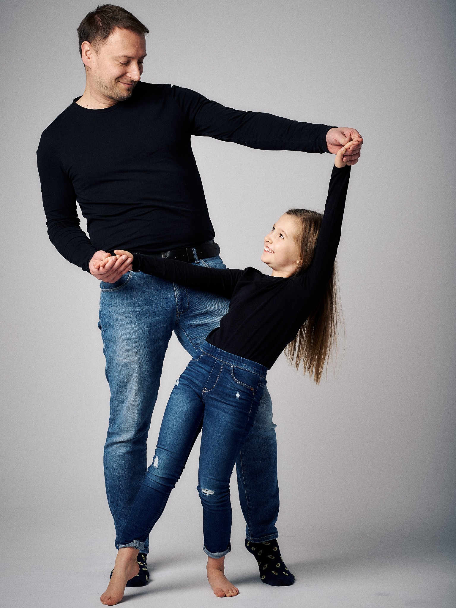 Ojciec w czarnej koszulce i jeansach trzyma za ręce uśmiechniętą córkę w podobnym stroju, która odchyla się do tyłu na szarym tle.