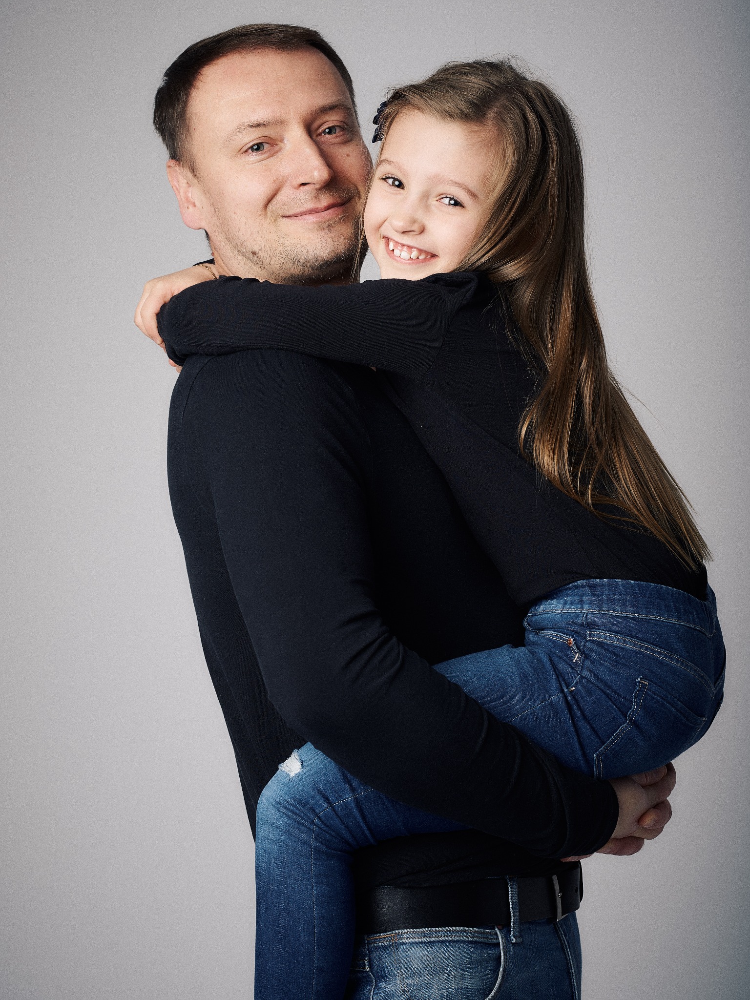 Uśmiechnięty tata trzyma na rękach córkę w jeansach, oboje ubrani na czarno, na jednolitym szarym tle.