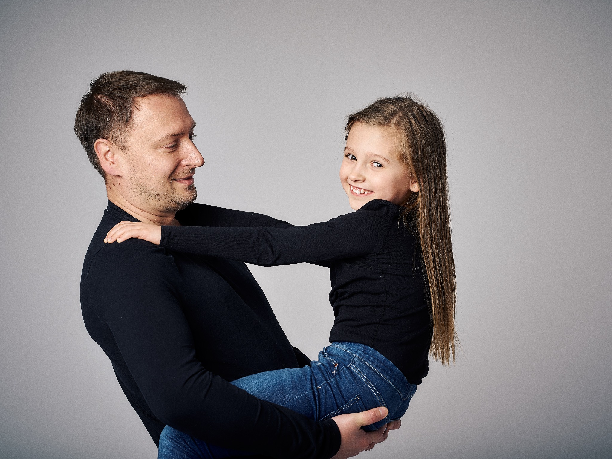 Uśmiechnięta córka w dżinsach obejmuje ojca, który trzyma ją na rękach na neutralnym tle w studiu fotograficznym.