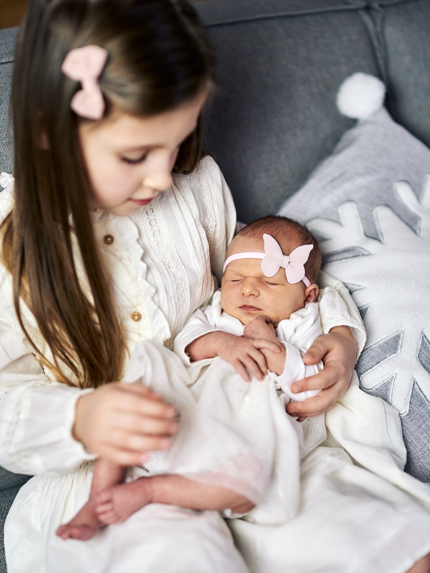 Starsza siostra w koronkowej sukience trzyma na kolanach śpiącą nowo narodzoną dziewczynkę z różową opaską w kształcie motyla na tle poduszki w kształcie śnieżynki.