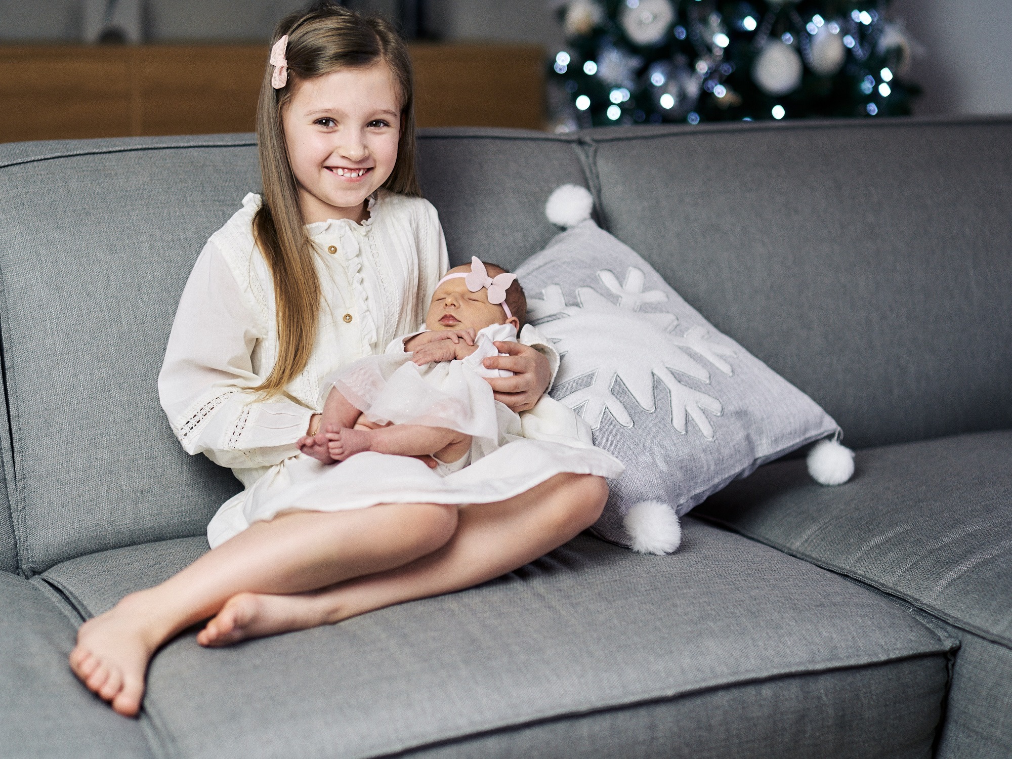 Uśmiechnięta dziewczynka trzyma na kolanach śpiące niemowlę z różową opaską na głowie, siedząc na szarym fotelu obok poduszki w kształcie śnieżynki, w tle rozmyte światełka choinkowe.
