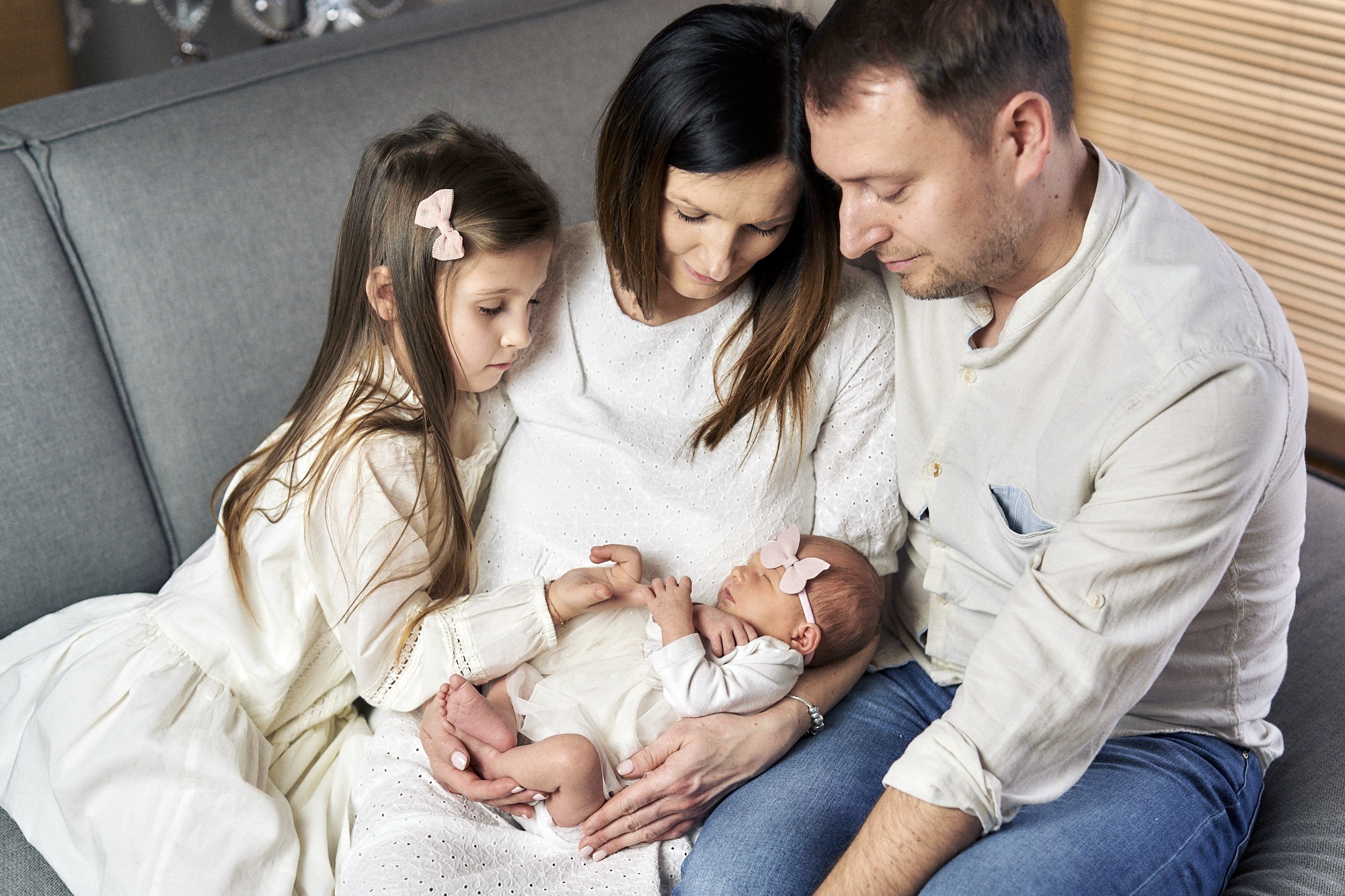Rodzina, w tym noworodek z różową kokardką na głowie, siedzi razem na szarej kanapie i patrzy na dziecko.