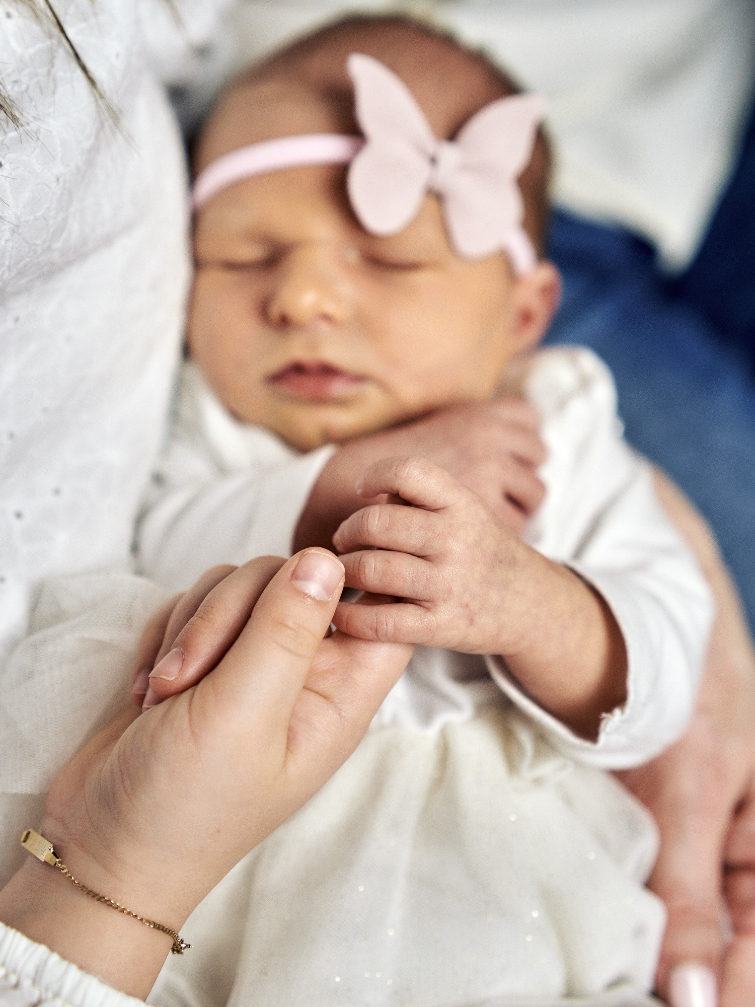 Śpiąca noworodka z różową opaską w kształcie motyla, trzymana w ramionach osoby z delikatną złotą bransoletką na nadgarstku.